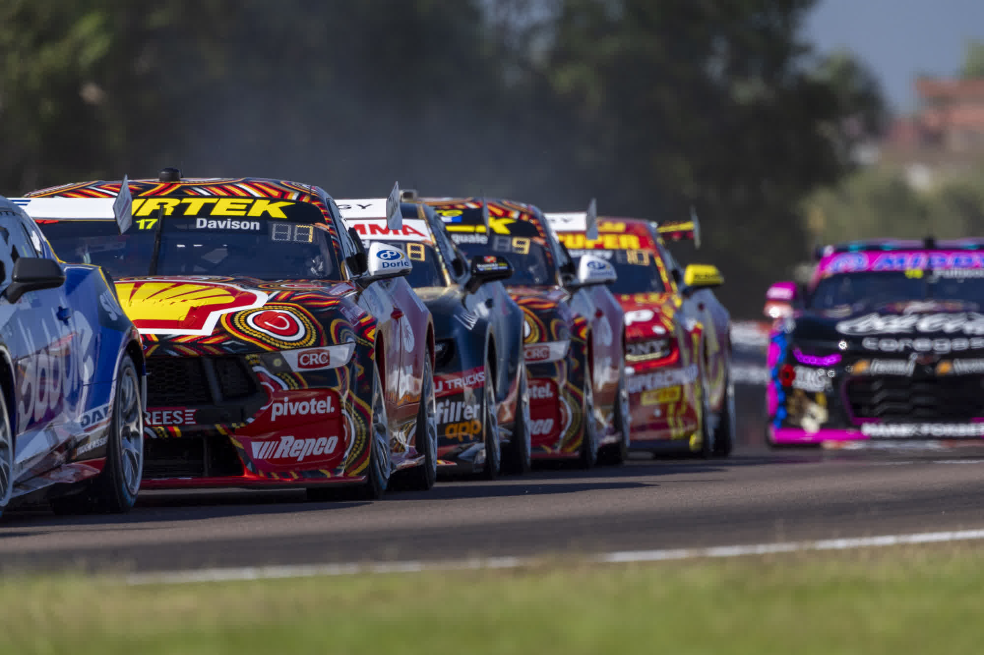 Race cars in vibrant colors speed down a track, closely packed, with blurred trees in the background on a sunny day.