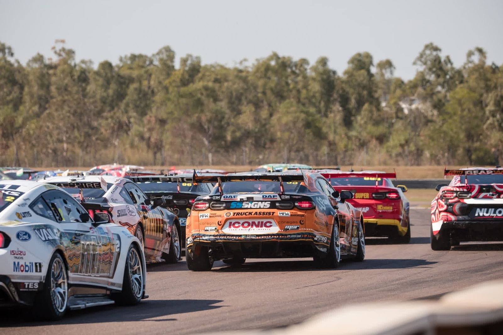 Race cars speed down a track, surrounded by trees. The vehicles display various sponsor logos and vibrant colors.